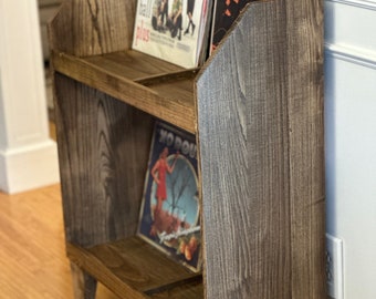 May include: A wooden record storage unit with two shelves, holding vinyl records. The unit is stained a dark brown with visible wood grain. The records have colorful album art, including one with the text "Gimme Gimmes".