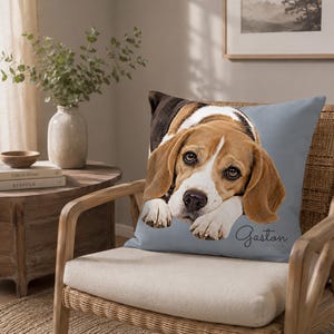 May include: A square throw pillow with a light blue background featuring a realistic portrait of a beagle dog resting its head on its front paws. The name Gaston is printed in a dark cursive script in the bottom right corner of the pillow.