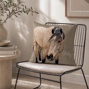 May include: A square throw pillow in a muted taupe color featuring a detailed illustration of a light tan horse with a dark mane and tail. The name Ritz is written in a white cursive script on the right side of the pillow cover.