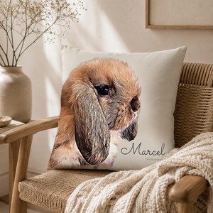 May include: A square throw pillow featuring a printed illustration of a brown lop-eared rabbit. The rabbit has long, soft ears and dark eyes. The name Marcel is printed in a cursive script font below the rabbit illustration on the off-white fabric.