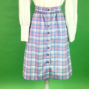 May include: A white long-sleeved blouse with a lace trim neckline and a plaid skirt with pink, blue, and white squares. The skirt has a button front closure.