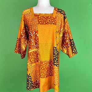 May include: A patchwork tunic in shades of orange, yellow, and brown, displayed on a white mannequin. The tunic features a square neckline and short sleeves. The fabric is patterned with various geometric and floral designs.