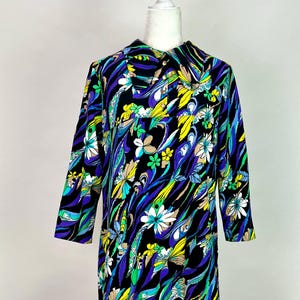 May include: A knee-length dress featuring a vibrant, multi-colored floral and abstract pattern on a black background. The garment has three-quarter length sleeves, a pointed collar, and a button-down front placket extending from the neckline.