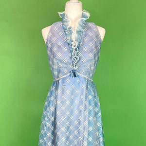 May include: A vintage blue and white plaid halter dress on a mannequin. The dress features a ruffled neckline, a fitted bodice, and a flowing skirt. The dress has a delicate white trim and a small tassel detail at the waist.