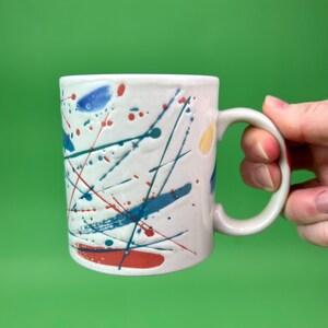 May include: A white ceramic mug with a colorful abstract design. The design features blue, red, and yellow paint splatters and lines.