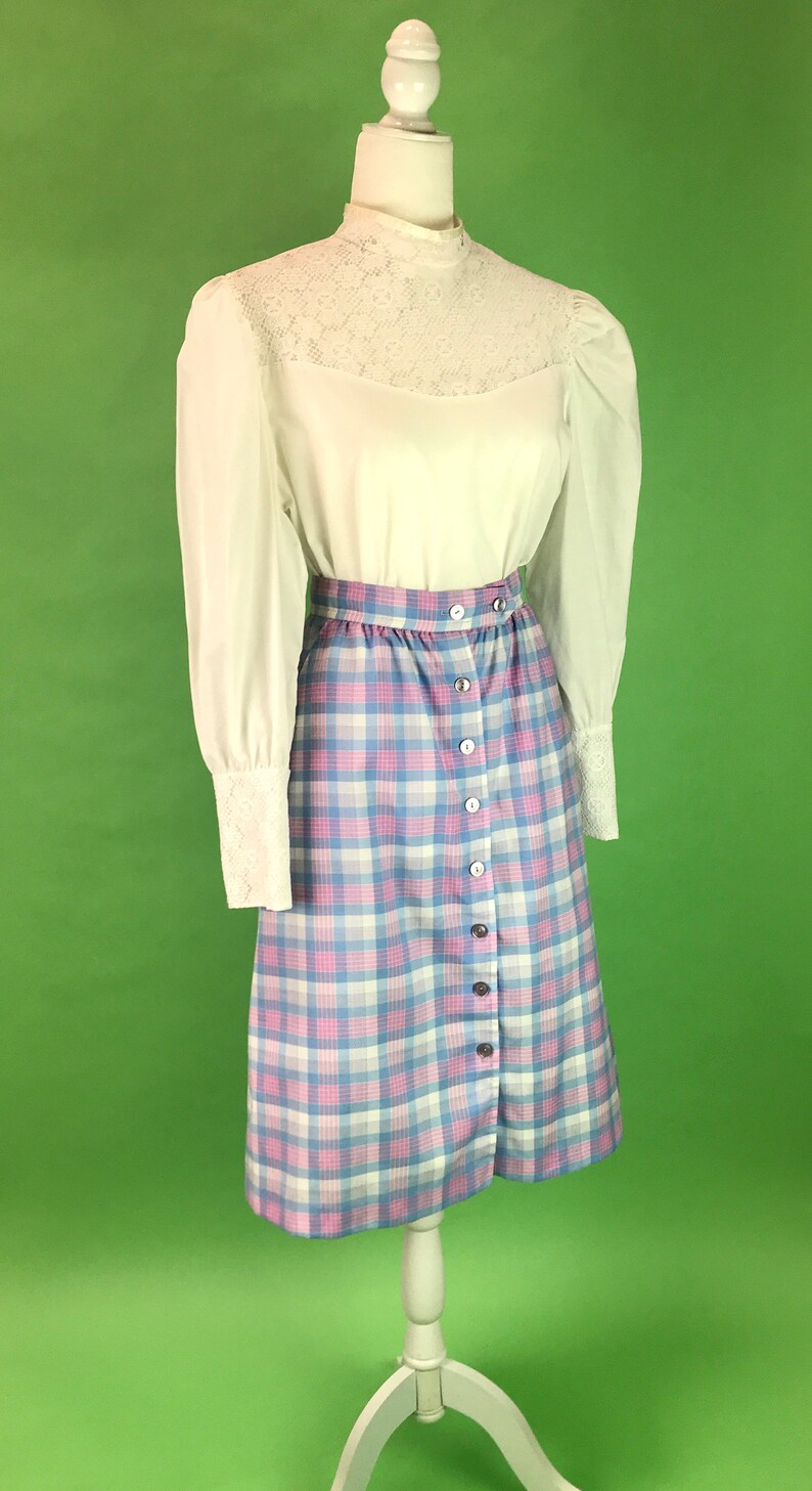 Op de afbeelding: Een wit blouse met een kanten kraag en lange mouwen, gecombineerd met een geruite rok in tinten roze, blauw en wit. De rok heeft een knoopsluiting aan de voorkant.