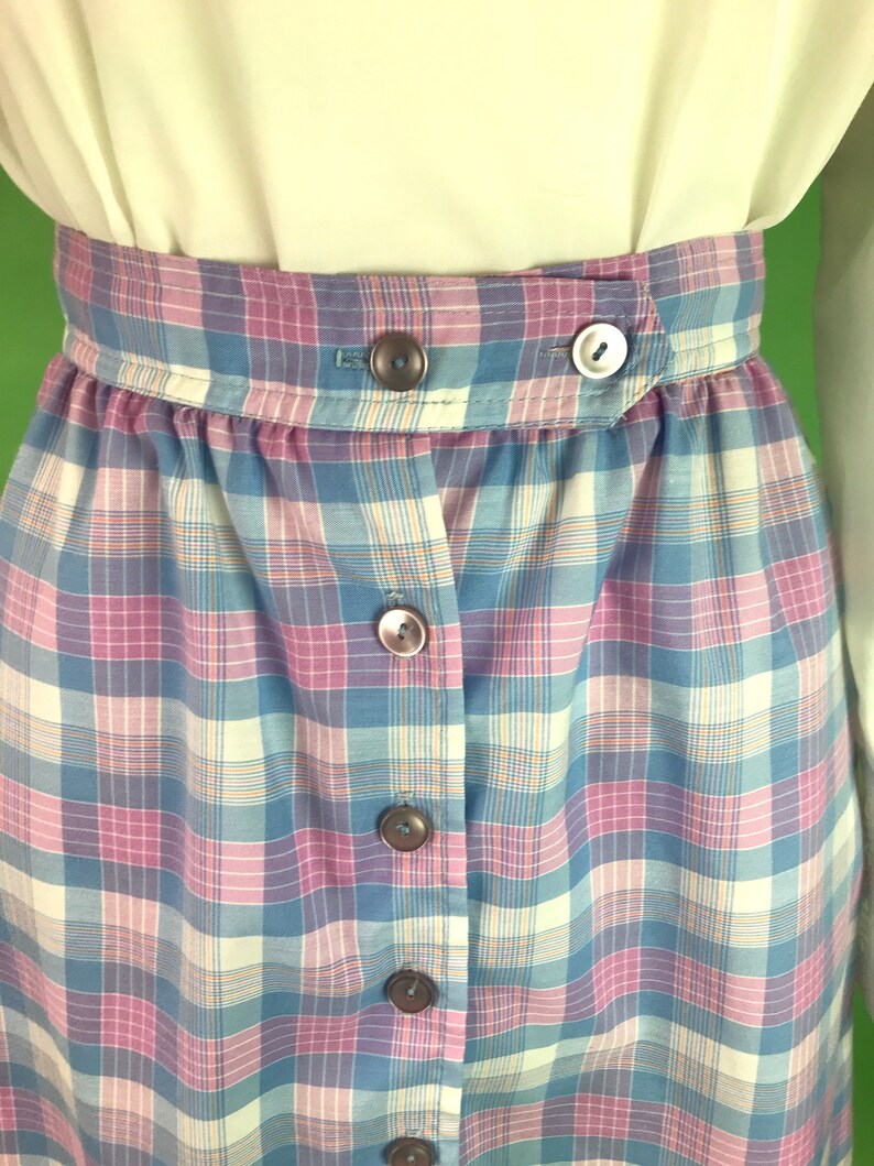 Op de afbeelding: Een blauwe, roze en witte geruite rok met knoopsluiting. De rok heeft een hoge taille en is gemaakt van een lichtgewicht stof.