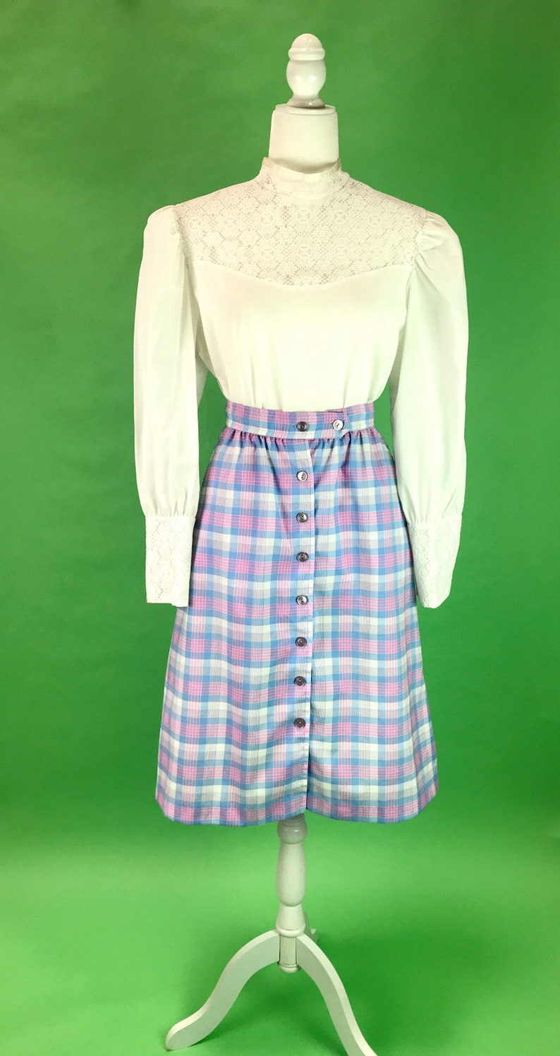 May include: A white long-sleeved blouse with a lace trim neckline and a plaid skirt with pink, blue, and white squares. The skirt has a button front closure.