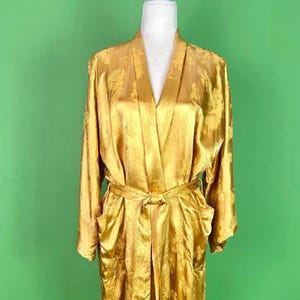 May include: A golden-yellow satin robe with a floral pattern, displayed on a mannequin. The robe has long sleeves, a shawl collar, a waist tie, and two front pockets. The background is a solid green.