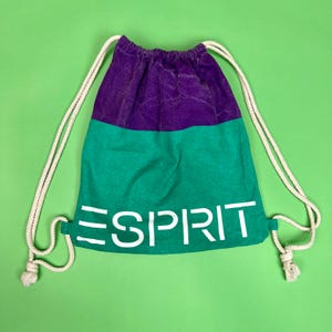 Puede incluir: Mochila con cordón de diseño de bloques de color. La sección superior es morada, la del medio es verde y la palabra "ESPRIT" está impresa en blanco. Tiene correas de cuerda blancas.
