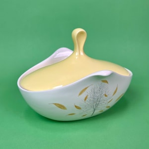May include: A white ceramic serving dish with a yellow lid and a gold leaf design. The dish is oval shaped and has a unique, modern design.