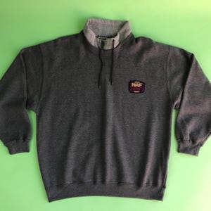 Op de afbeelding: Een donkergrijze sweatshirt met een hoge kraag en een kleine logopatch. De kraag is lichtgrijs en de sweatshirt heeft een trekkoord bij de hals. De logopatch leest "NAF" in witte letters op een bordeauxrode en zwarte achtergrond.