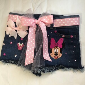 May include: Blue denim shorts with a pink and white polka dot bow and a pink Minnie Mouse applique. The shorts are decorated with rhinestones.