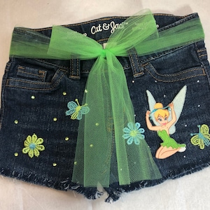 May include: Blue denim shorts with a green tulle bow, embellished with green and blue flowers, butterflies, and a Tinkerbell applique. The shorts have a frayed hem.