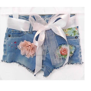 May include: Blue denim shorts with a white satin ribbon tied around the waist. The shorts have a floral print and a pink flower embellishment.
