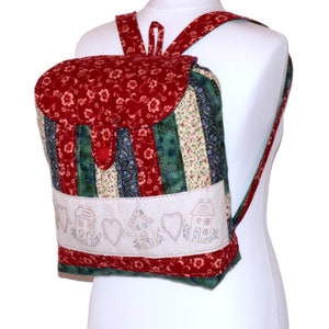 Op de afbeelding: Een rode, groene en witte patchwork stoffen rugzak met een klep sluiting en een klein geborduurd ontwerp van huizen en harten.