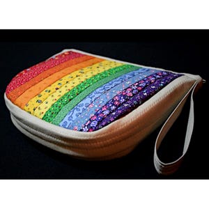 Może przedstawiać: Ręcznie robiona kopertówka z tęczowym wzorem. Torba posiada paski z czerwonej, pomarańczowej, żółtej, zielonej, niebieskiej i fioletowej tkaniny. Posiada kremowy zamek błyskawiczny i pasek na nadgarstek. Tło jest czarne.