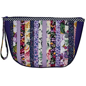 Może przedstawiać: Pikowana kopertówka z kwiatowym wzorem patchworkowym. Torba posiada pionowe paski materiału w różnych kwiatowych wzorach, lawendowym i granatowym. Czarno-biały wzór w kratkę zdobi górną krawędź, a fioletowy pasek na nadgarstek jest przymocowany.