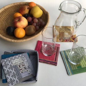 Peut inclure: Quatre dessous-de-verre en tissu coloré avec un verre d'eau sur un dessous-de-verre vert et un pichet en verre sur un dessous-de-verre fleuri. Un bol de fruits repose sur un panier en osier.