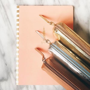 May include: Three pencil cases in different colors and materials. The top case is gold with a zipper closure. The middle case is silver with a blue zipper. The bottom case is brown leather with a zipper closure.