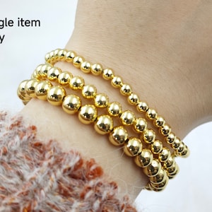 May include: Three gold beaded bracelets stacked on a person's wrist. The bracelets are made of round, shiny gold beads.