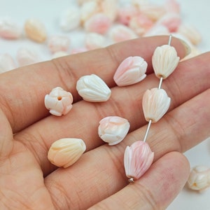 Puede incluir: Primer plano de una mano sosteniendo una hebra de cuentas de flores de concha rosa y blanca. Las cuentas están talladas en forma de flor con una base redondeada y una parte superior puntiaguda. Las cuentas están enhebradas en un alambre plateado.