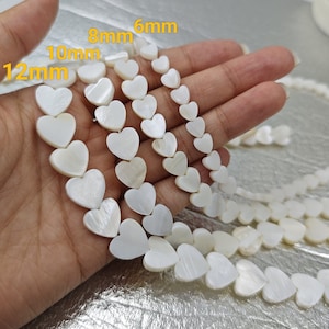 Peut inclure: Un brin de perles de coquillage en forme de cœur blanches en quatre tailles différentes : 6 mm, 8 mm, 10 mm et 12 mm. Les perles sont enfilées sur un cordon blanc et sont présentées sur une main.