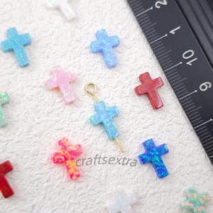 Puede incluir: Una colección de pequeños dijes en forma de cruz de varios colores, incluyendo azul, rosa, rojo y blanco iridiscente, dispuestos junto a una regla negra con medidas en centímetros. Un dije de cruz azul incluye un perno de ojo dorado.