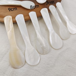 May include: Seven white mother of pearl spoons of varying sizes. The spoons are arranged in a row on a white surface. The spoons are 7 cm, 8 cm, and 9 cm in length.