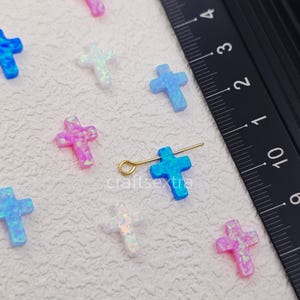 May include: A collection of small cross-shaped charms in blue, pink, and white opal-like material. One blue cross is attached to a gold-toned metal eye pin. A black ruler is visible on the right, showing measurements in centimeters.