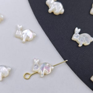 Puede incluir: Una colección de dijes en forma de conejo blanco iridiscente, algunos con un pasador dorado. Los dijes están dispuestos sobre una superficie blanca y negra.