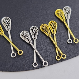 Pode incluir: Uma coleção de pingentes de metal em forma de tacos de lacrosse em ouro e prata. Os pingentes têm um design de rede detalhado na cabeça e uma argola na extremidade do cabo. Dispostos sobre uma superfície escura.