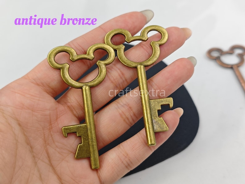 May include: Two antique bronze keys with a rounded, mouse-ear shaped bow at the top. The keys feature a straight shank and a notched bit at the bottom. The text "antique bronze" and "craftextra" are visible in the image, along with a third key.