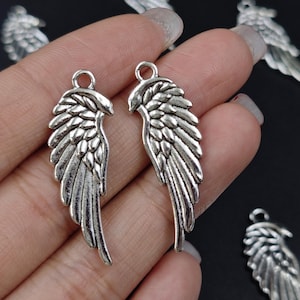Op de afbeelding: Twee zilveren engelenvleugelbedels met een gedetailleerd verenontwerp. De vleugels zijn ongeveer 5 cm lang en hebben een lus aan de bovenkant om aan sieraden of andere knutselwerken te bevestigen.