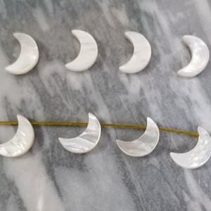 Puede incluir: Ocho cuentas blancas en forma de media luna hechas de madreperla. Las cuentas están dispuestas en dos filas sobre una superficie de mármol.