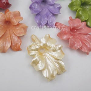 Op de afbeelding: Een verzameling veelkleurige bloemvormige kralen met een glanzende, parelmoerachtige afwerking. Het assortiment bevat individuele stukken in de kleuren perzik, lavendel, limoengroen, crème, rood en blauw, elk met een centraal gat.