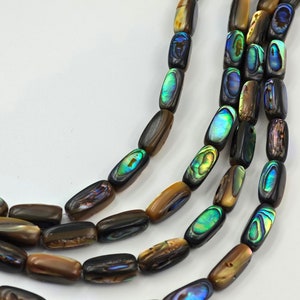 Puede incluir: Una hebra de cuentas de concha de abulón iridiscente de forma rectangular. Las cuentas son una mezcla de marrón, verde y azul.