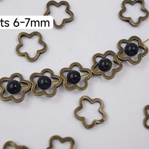 Puede incluir: Espaciadores de cuentas en forma de flor de color bronce con cuentas oscuras ensartadas en un alambre dorado. El texto "Fits 6-7mm" está en un cuadro blanco. Varias cuentas de flores sueltas están dispersas.