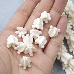 May include: A handful of small, off-white, flower-shaped beads. The beads have a detailed, textured design and are scattered across a hand. A strand of similar beads is visible on the right side. The background is a light blue.