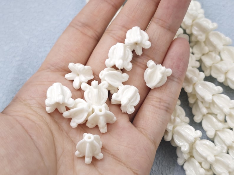 May include: A handful of small, off-white, flower-shaped beads. The beads have a detailed, textured design and are scattered across a hand. A strand of similar beads is visible on the right side. The background is a light blue.