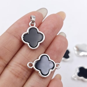 Peut inclure: Charmes trèfle à quatre feuilles avec incrustations d'onyx noir et sertis en métal argenté. Les charmes sont présentés sur une main et sur une surface blanche.