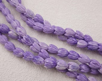 May include: Close-up of several strands of lavender-colored beads. Each bead is shaped like a flower bud with detailed ridges. The beads are arranged diagonally across a textured, off-white surface, creating a visually appealing pattern.