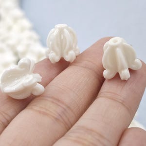 May include: Close-up of three small, white, flower-shaped beads. Each bead has a detailed, petal-like design and a small hole at the top. The beads are held in a hand, with a blurred background.
