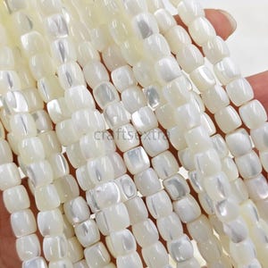 Peut inclure: Une collection de perles en nacre blanche disposées en plusieurs brins verticaux. Chaque perle présente une forme rectangulaire arrondie et lisse avec un éclat irisé naturel et une finition polie, reflétant la lumière sur sa surface.