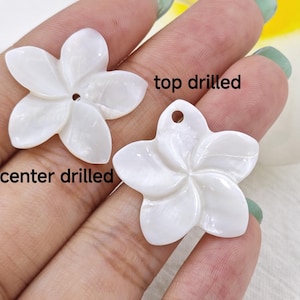 Op de afbeelding: Twee witte schelpbloem bedels, een met een gat in het midden en de andere met een gat aan de bovenkant. De tekst "center drilled" staat onder de eerste bedel en "top drilled" staat boven de tweede bedel.