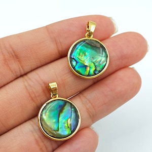 Könnte beinhalten: Zwei runde Anhänger mit irisierenden grün-blauen Abalone-Muscheln, gefasst in goldfarbenem Metall. Jeder Anhänger hat oben eine kleine Schlaufe für eine Kette. Die Anhänger werden vor weißem Hintergrund gezeigt.
