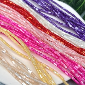 May include: Close-up of several strands of colorful beads in shades of pink, red, purple, white, and yellow. The beads are small, rectangular, and strung together, creating a vibrant and textured display. The beads are arranged on a white surface.