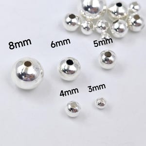 May include: Silver beads in various sizes, from 3mm to 8mm, arranged on a white surface. The beads are round and have a smooth, shiny finish.