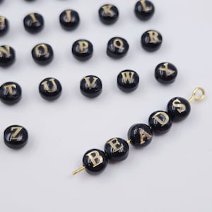 Puede incluir: Cuentas de alfabeto negras con letras doradas que deletrean la palabra "BEADS". Las cuentas están enhebradas en un alambre dorado con un bucle dorado en el extremo.