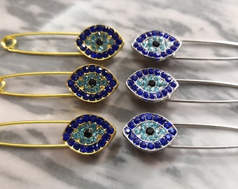Puede incluir: Seis imperdibles con dijes de ojo turco azul y turquesa. Los dijes están hechos de pedrería y tienen un centro negro. Tres imperdibles son de metal dorado y tres de metal plateado.
